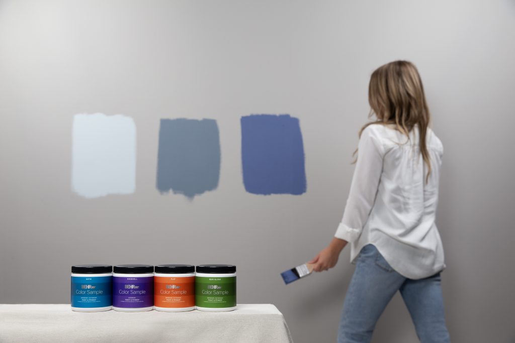 A woman testing out paint samples on the wall. She has three paint swatches painted on the wall each 6 inches apart from one another. In the foreground is the paint samples jars. 