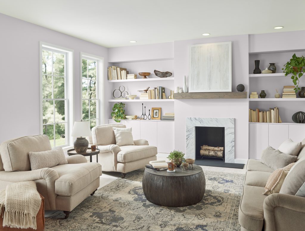 A bright and welcoming living room with an almost neutral color palette. The room has large windows curtains, allowing natural light to fill the space. A fireplace is present on the right side of the image, above which hangs an abstract piece of art.