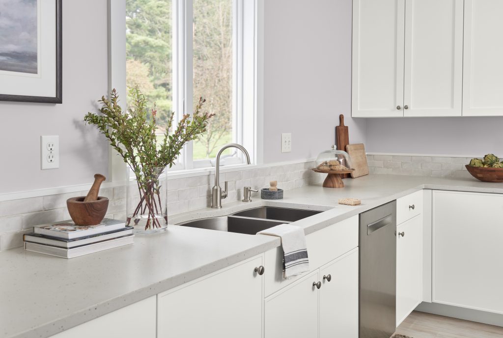 A kitchen with light gray-violet color walls and white cabinetry features a white countertop with a built-in stainless-steel sink. The counter includes a wooden mortar and pestle, a stack of books, and a vase with green foliage. To the right of the sink, there are cutting boards, a cake stand with pastries, and a bowl of fruit. Large windows allow natural light to fill the space.