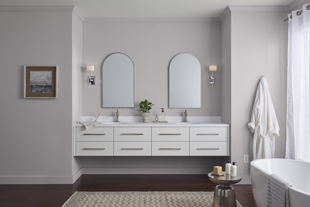 A modern bathroom features a double vanity with two mirrors, light fixtures, and sinks. A small plant sits between the sinks. To the right, a white robe hangs on the light purple-gray wall near a freestanding bathtub with candles and decorative items on its edge. The floor is dark wood, with a light-colored rug in front of the vanity.