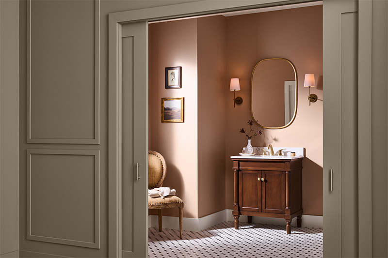 An elegant bathroom with warm light warm, pink-colored walls, a wooden vanity with white countertop, and a gold-framed oval mirror. Two wall sconces with peach shades flank the mirror, and a small chair with folded towels sits against the side wall beneath framed artwork.