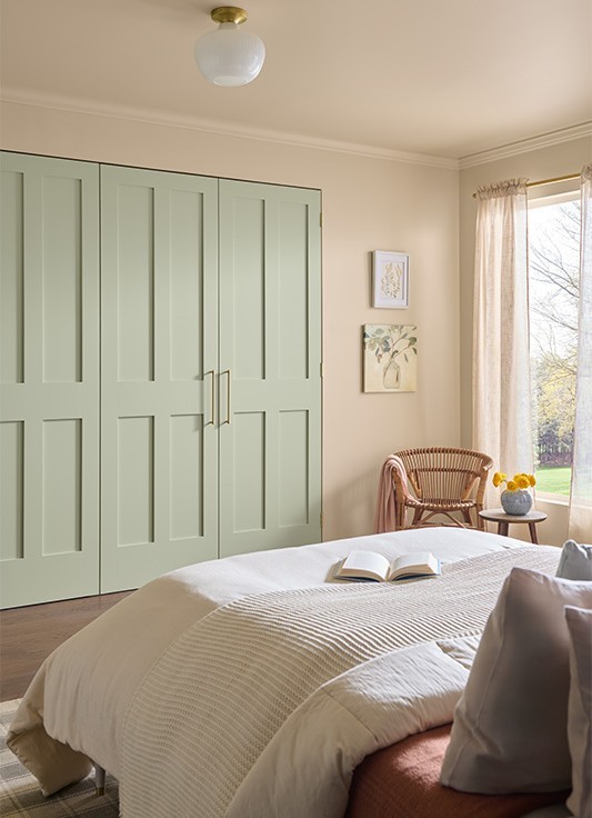 A casual cozy bedroom featuring a bed with a beige comforter and an open book. Natural light streams in through a large window with light curtains. Beside the window is a small round table with a vase of yellow flowers and a wicker armchair. The back wall displays two framed artworks, and the left side of the room has large green closet doors with gold handles. 
