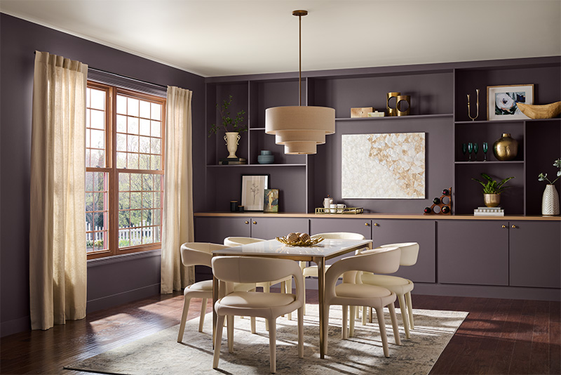 An eclectic dining room with dark purple walls and a large window with beige curtains. The room features a white dining table surrounded by off-white chairs on a light-colored rug. Above the table hangs a beige, tiered pendant light. The back wall has built-in shelves and cabinets, decorated with various items including vases, books, plants, and artwork.