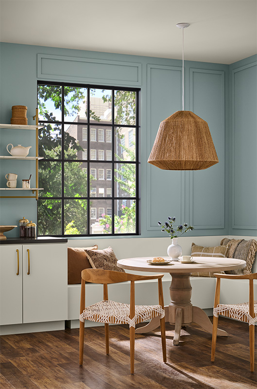 A bright dining nook with soft blue-green walls and a large, black-framed window. A round wooden pedestal table is surrounded by woven-seat chairs and a built-in bench with neutral throw pillows.