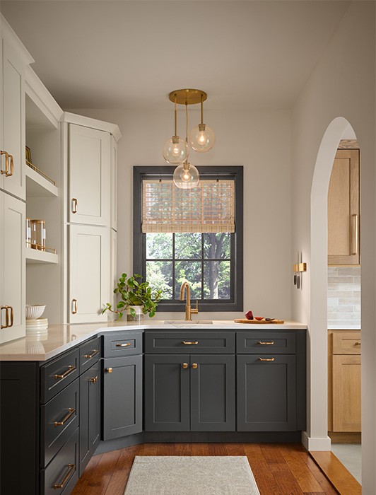 A beautiful kitchen pantry with two-tone cabinetry featuring dark gray lower cabinets and light beige upper cabinets, accented with brass hardware. A black-framed window above the sink has woven blinds, with a brass faucet below.