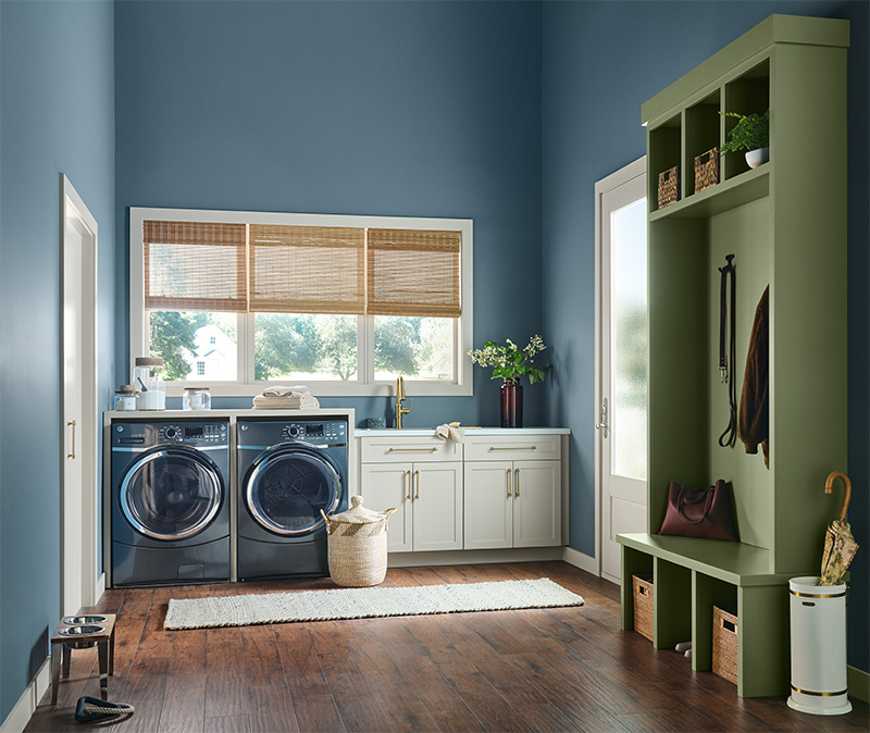 A laundry room with blue walls and wooden flooring. It features a front-loading washer and dryer, a white countertop with lower cabinets, and a window with bamboo blinds above. On the right is a green built-in storage unit with cubbies, hooks, and a bench with storage baskets. The room is decorated with household items like plants, jars, and cleaning supplies. 