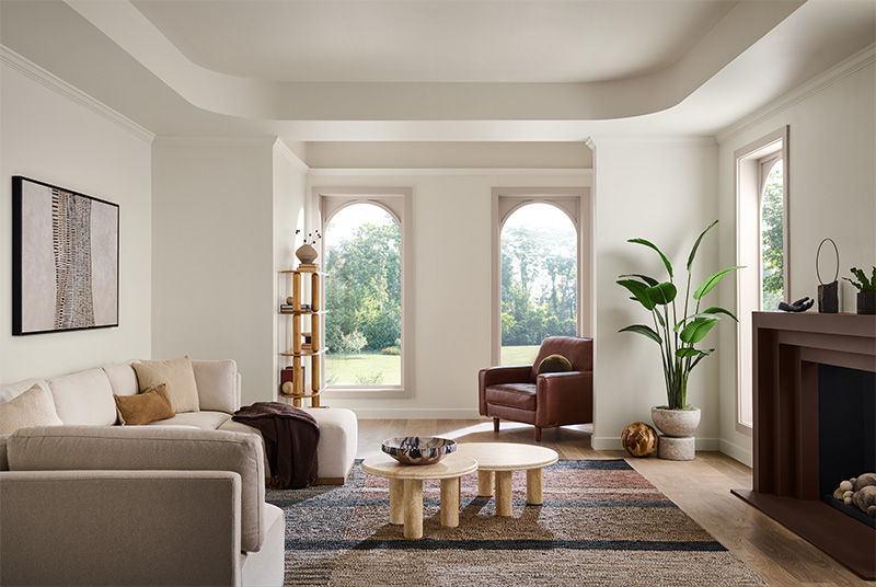 A modern living room with large arched windows overlooking greenery. The space features a beige sofa with cushions, a brown leather armchair, and nested round coffee tables. A tall potted plant stands beside a fireplace, with a smaller plant on the mantel. A wooden shelving unit with decorative items sits near a window, and an abstract painting hangs above the sofa. The room has white walls and a patterned rug in neutral tones.