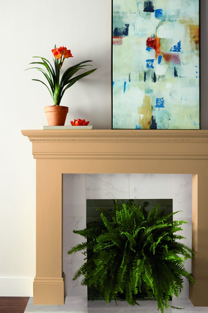 Beige-painted fireplace mantel with orange flowers, abstract art, and a green fern inside; white wall background.