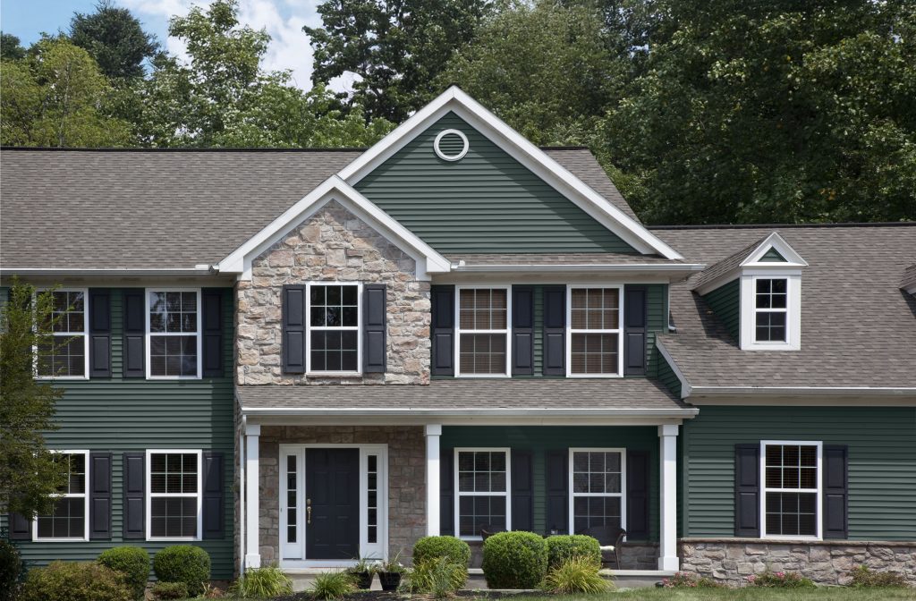 A two-story house with green siding and a stone facade at the entrance. Features a gabled roof with white trim, black shutters, and a dormer window. The front door is black and there are three white columns supporting the porch overhang. 