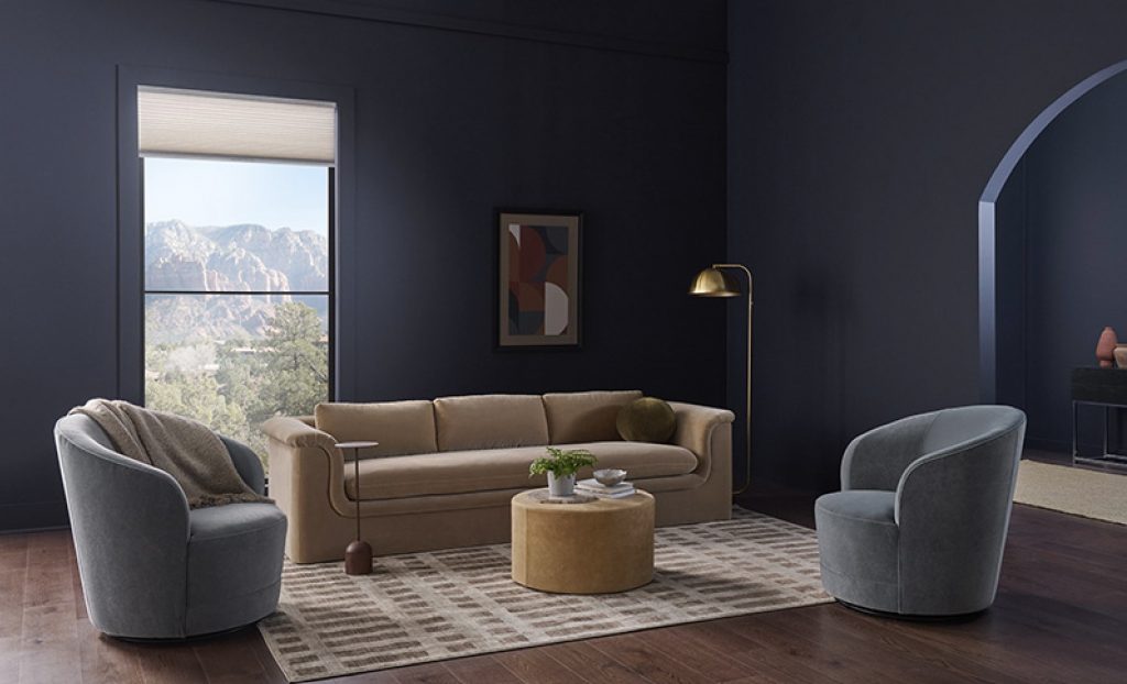 Image shows living room with dark blue walls, beige sofa, grey chairs, mustard ottoman, and a large window with mountain view.