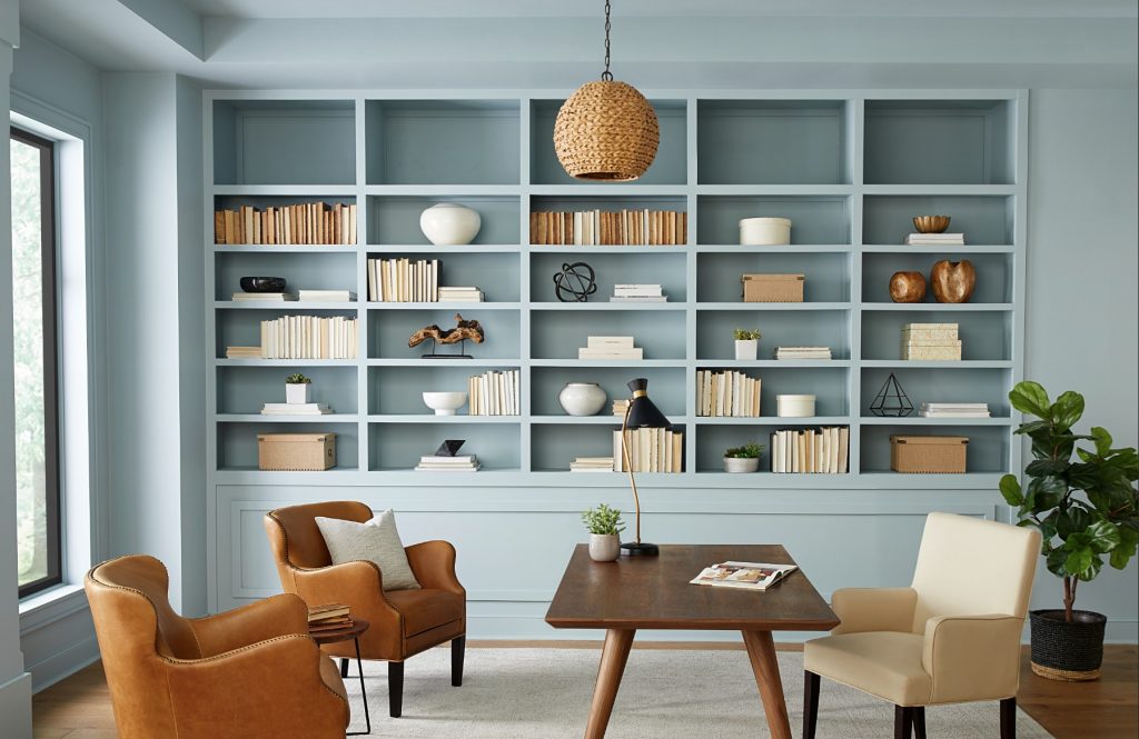 Home office with light blue built-in bookshelf, wooden desk, leather and upholstered chairs, and natural light from large windows.