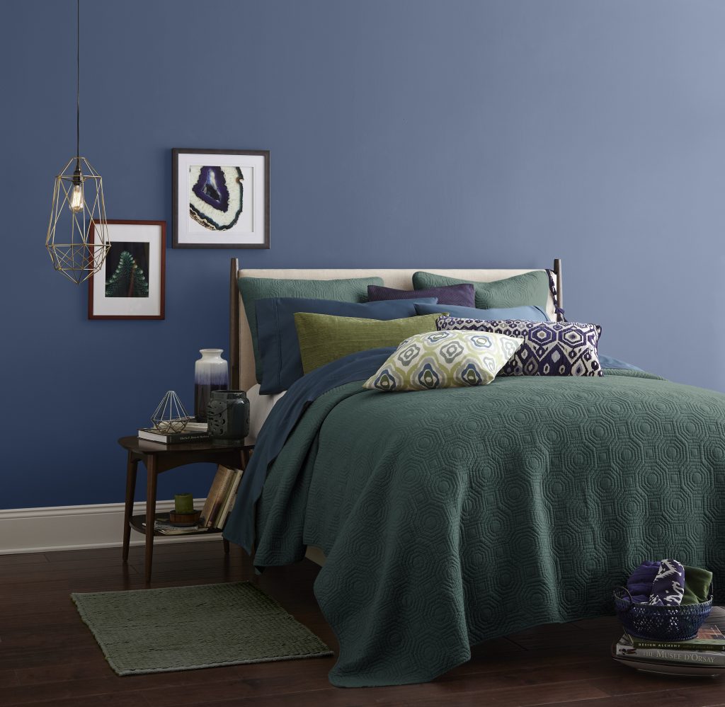 A bedroom with walls painted in a calming blue hue painted on the walls. Bedding is in green, blue and purple jewel tones.