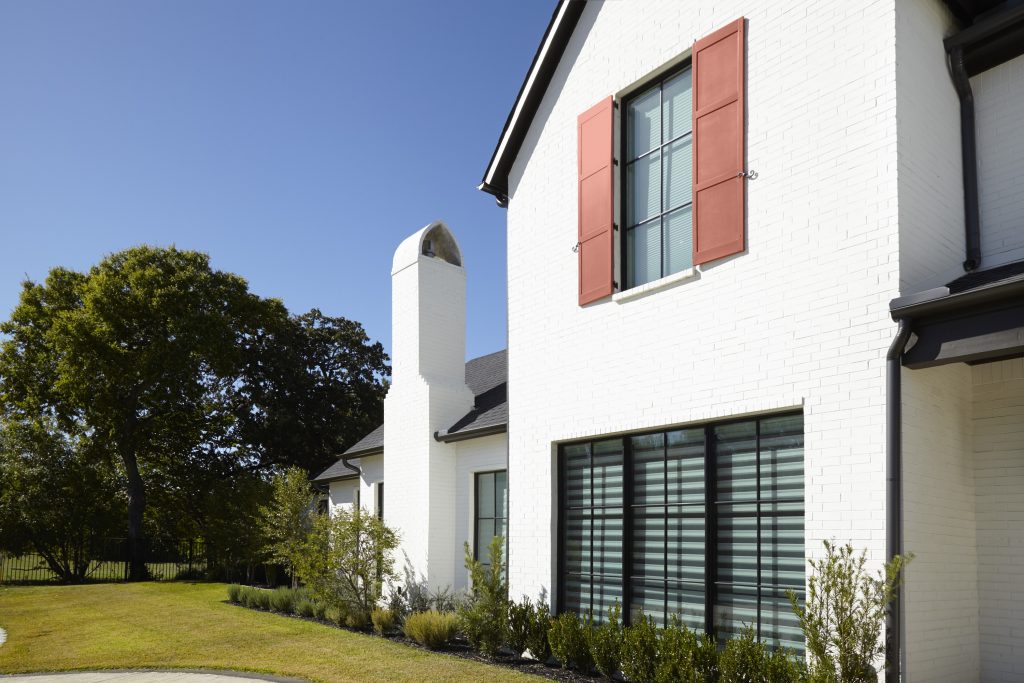 White brick house with black trim and a steep gabled roof. The upper window features terra cotta-colored shutters, and the home has a tall white chimney. The front yard includes green grass, shrubs, and a large tree under a clear blue sky.
