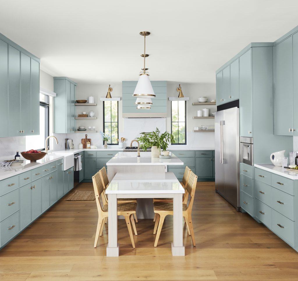 A spacious kitchen with cabinets painted in a soft blue-green tone called Watery. The room features a white island, a white eating area and light wood dining chairs. White colored backsplash and countertops and warm wood flooring contrasts beautifully with the cool-toned cabinetry. 