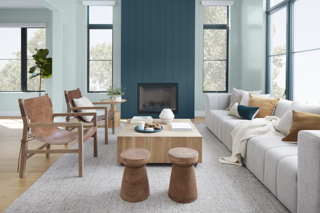 A modern living room with a lot of natural light and painted with Watery. It features a tall shiplap fireplace wall painted in a deep blue shade called Nocturne Blue. The space features a light gray sofa, a wooden armchair with a tan cushion, and a square wooden coffee table with decorative objects. 