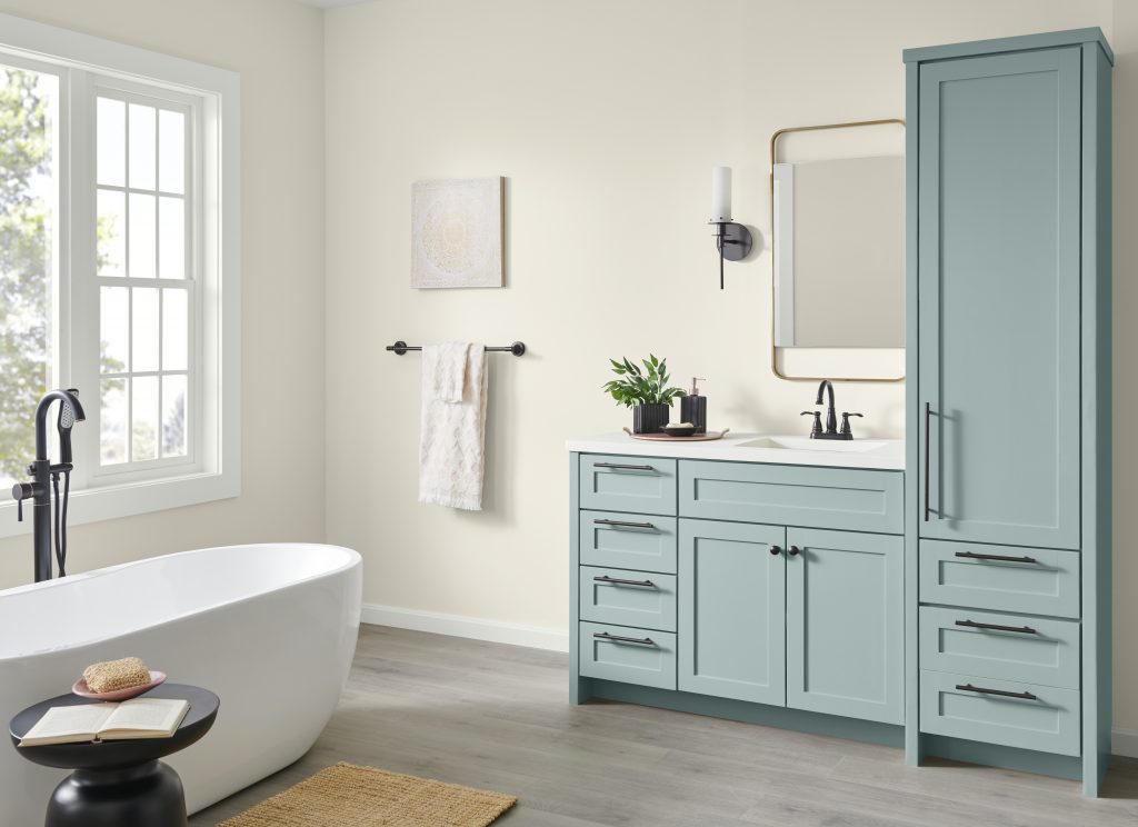 A spacious bathroom features a freestanding white tub by a large window and a vanity painted in a blue-green shade called Watery. The countertop is white, and a tall storage cabinet, black fixtures, a gold-framed mirror, and a modern sconce add style. Light neutral floors and a woven natural-material mat complete the serene, spa-like look.