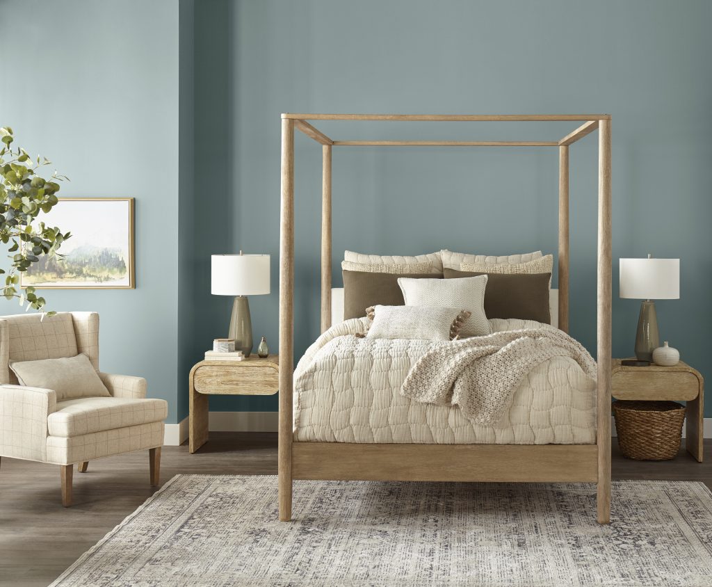 Cozy bedroom featuring a four-poster wooden bed with layered neutral bedding, including a quilted comforter and textured throw blanket. The walls are painted in a soft light blue, creating a calm and balanced atmosphere. Two wooden nightstands with lamps flank the bed, complemented by a light armchair, a patterned area rug and a potted plant for greenery.