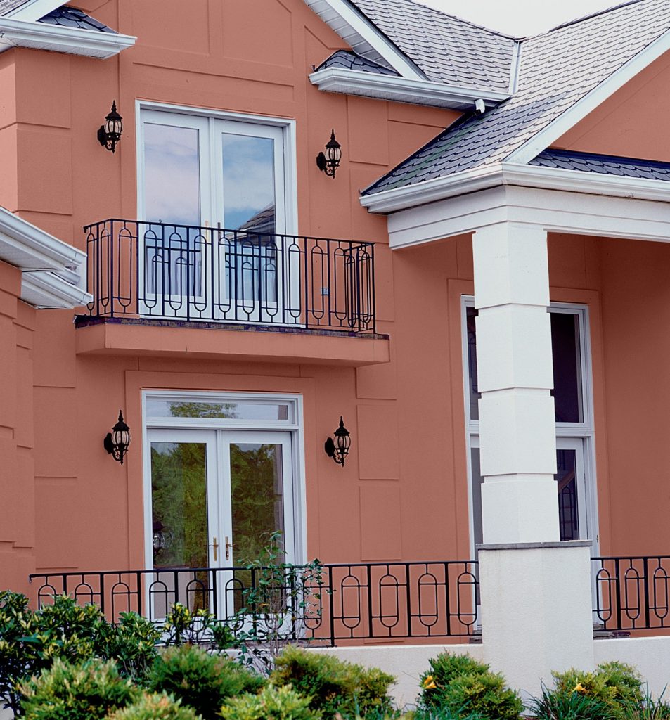 Close-up of a two-story home with exterior walls painted in a warm terra cotta color. The house has white columns, black wrought iron railings on balconies, and white-framed French doors. 