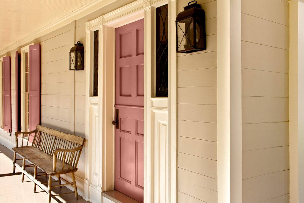 A traditional front porch featuring terra cotta color door and shutters against a soft beige exterior, accented with black lanterns and a wooden bench.