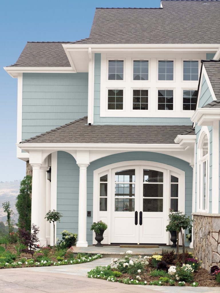 A two-story home exterior with soft blue siding, crisp white trim, and shingle roof, featuring a covered front porch with white columns, double front doors, and landscaped garden beds.

