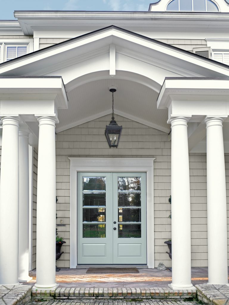 A covered front porch with white columns, light neutral shingle siding, and pale blue double doors beneath a hanging lantern.