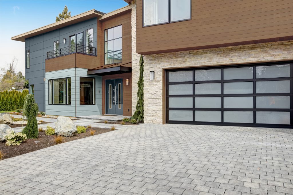 A modern two-story home with mixed exterior materials, including gray-blue, light dusty blue and wood-toned siding, stone accents, large windows, and a contemporary garage door with a paved driveway.

