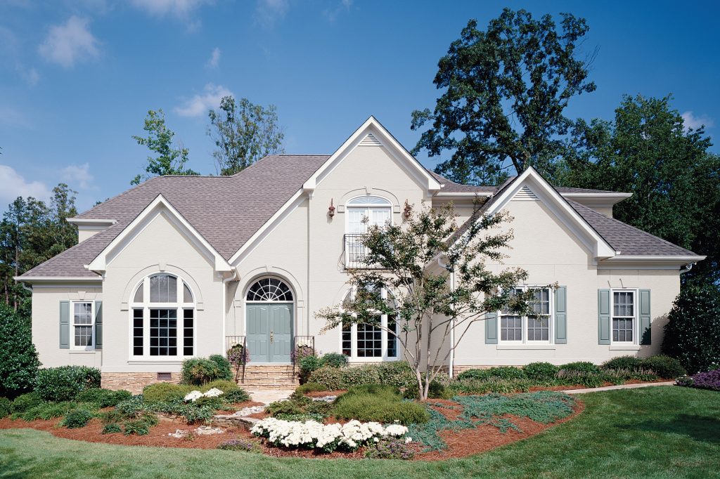 A large traditional home with a warm white exterior, blue shutters, an arched front entry, and manicured landscaping set against a blue sky.