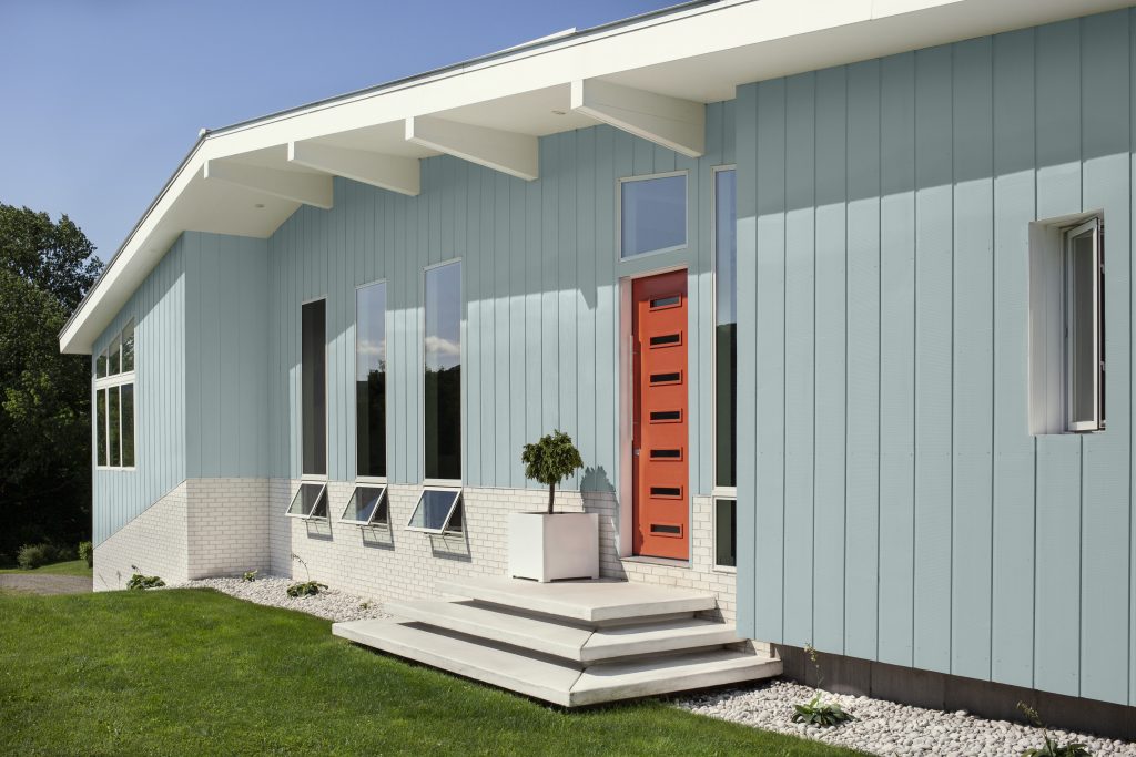 A mid-century modern home exterior with light blue vertical siding, a bold red-orange front door, white brick base, and a simple front stoop set against a green lawn.