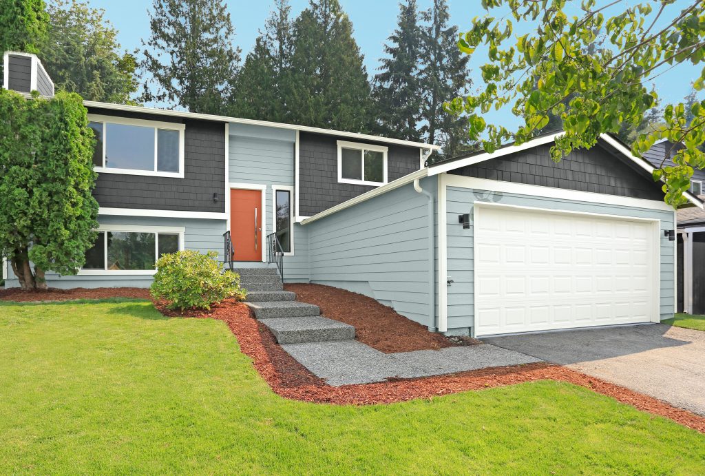 A Ranch, split-level home with dark gray and light blue siding, a white garage door, an orange front door, and a landscaped front yard with concrete steps.