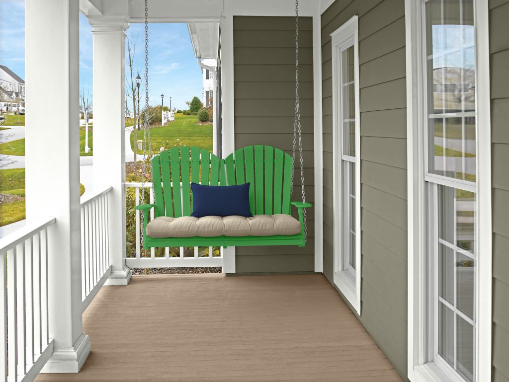 A front porch with white columns and railing, green porch swing with beige cushion and navy blue pillow, light wood flooring, and muted green color house siding overlooking a grassy neighborhood. The porch wood stain color is Taupe and the finish a semi-trasparecent opacity. 