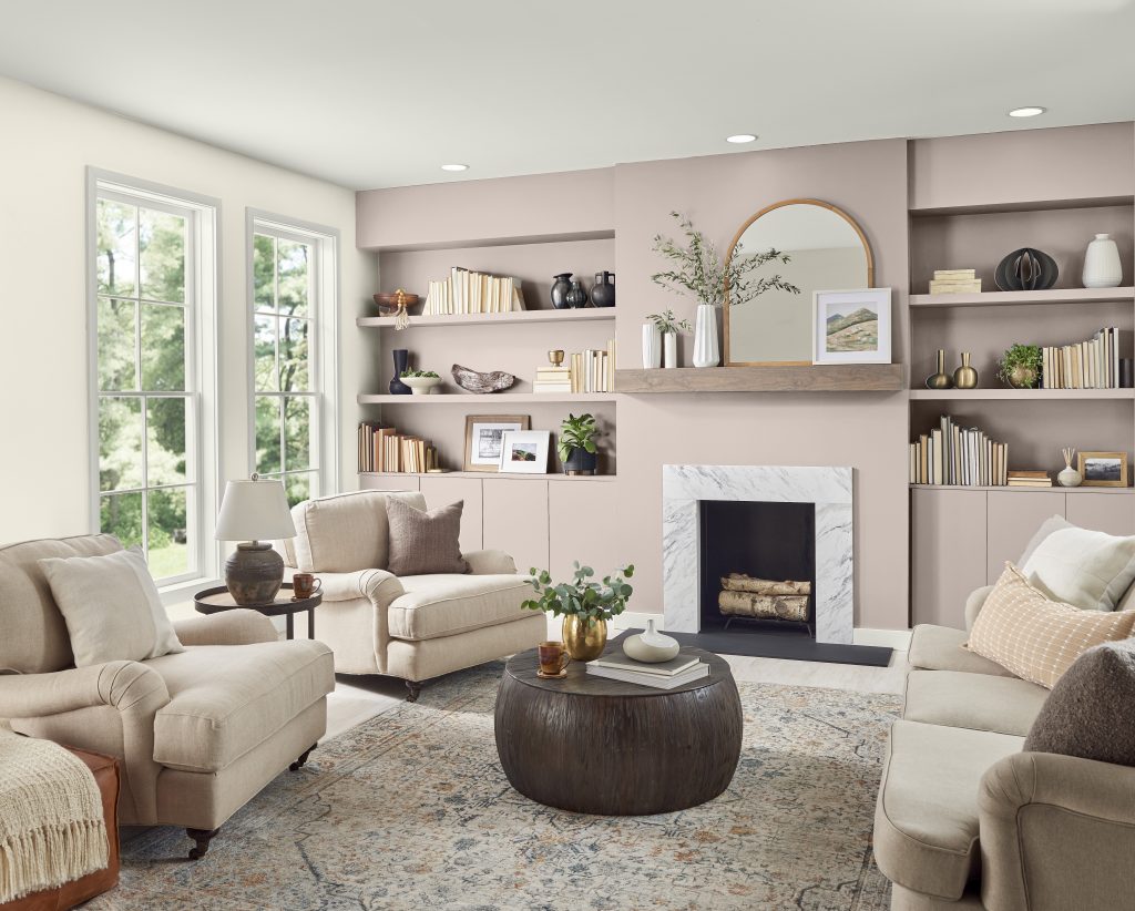 A cozy living room with soft blush pink walls and built-in shelving and white marble fireplace. The shelves are styled with books, vases, and small plants, and a round mirror hangs above the mantel. Neutral upholstered chairs and a sofa surround a large round wooden coffee table on a patterned area rug.