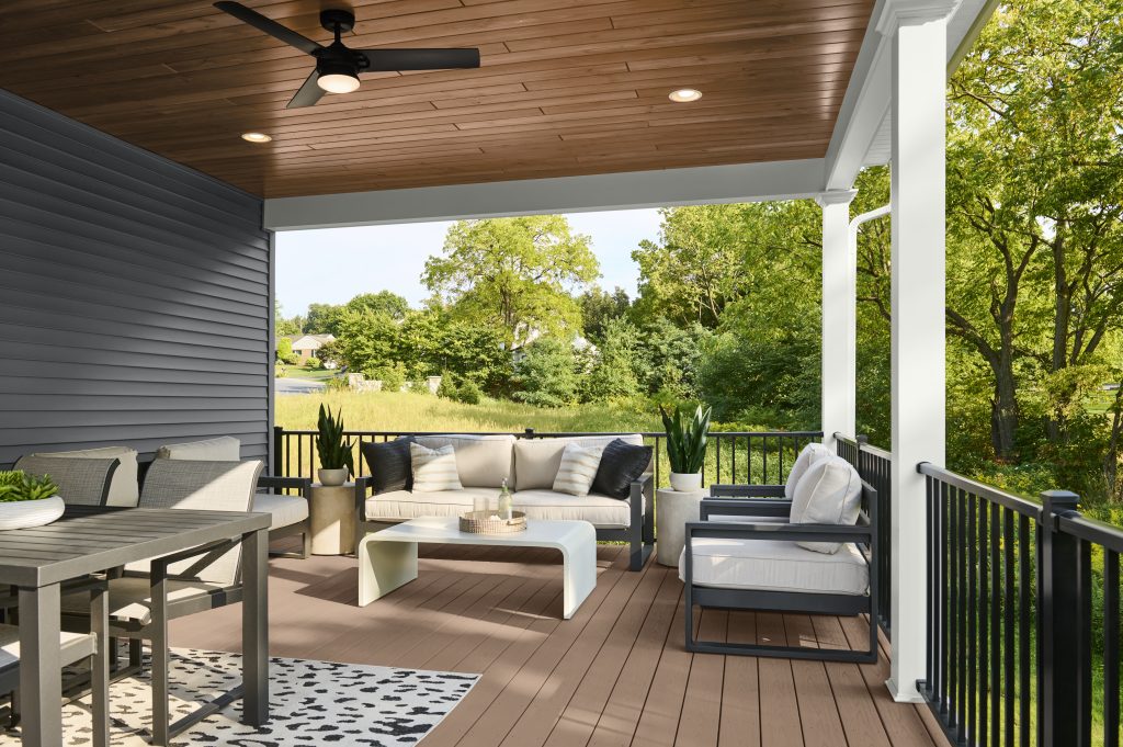 A covered porch with stained wood ceiling and matching stained deck boards, furnished with outdoor seating, a dining table, and a ceiling fan, overlooking a green landscape.