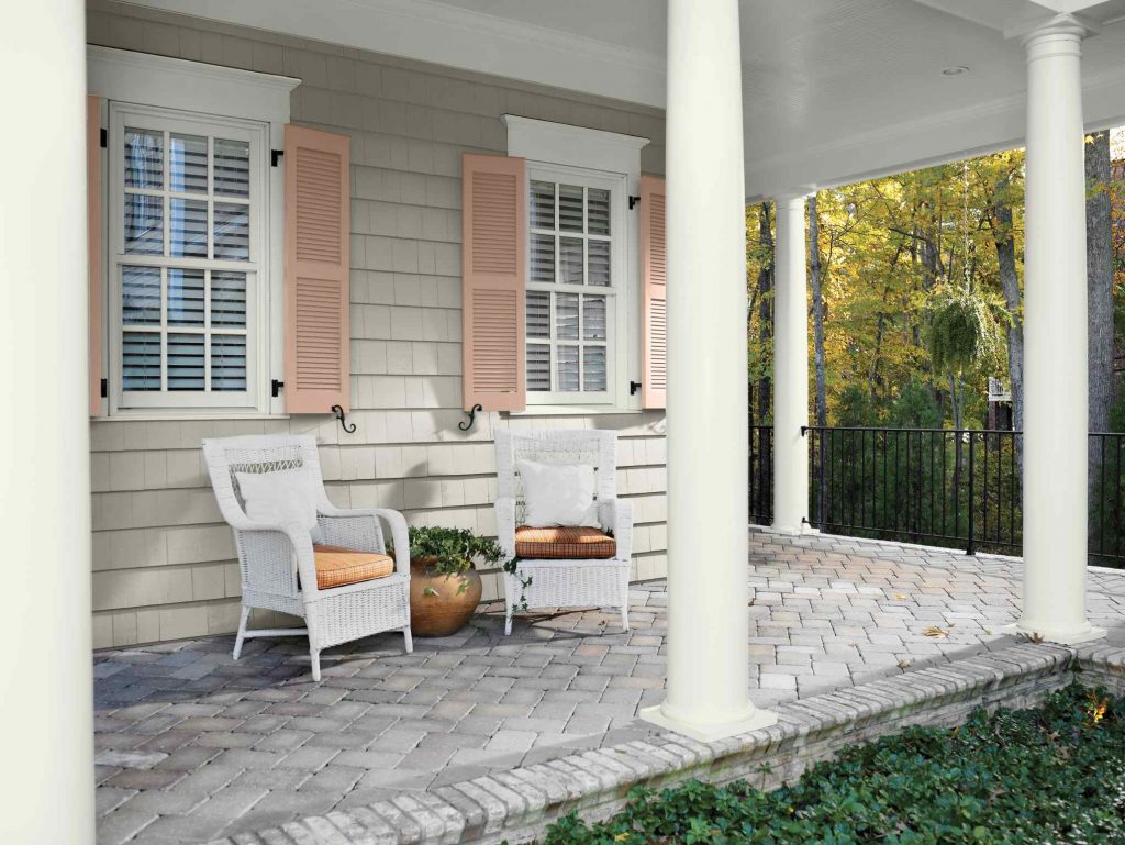 An inviting covered front porch with siding painted Gray View, crisp Polar Bear trim, and warm Iced Copper shutters, featuring two white wicker chairs on a brick paver floor, large white columns, and a peaceful garden view beyond the porch.