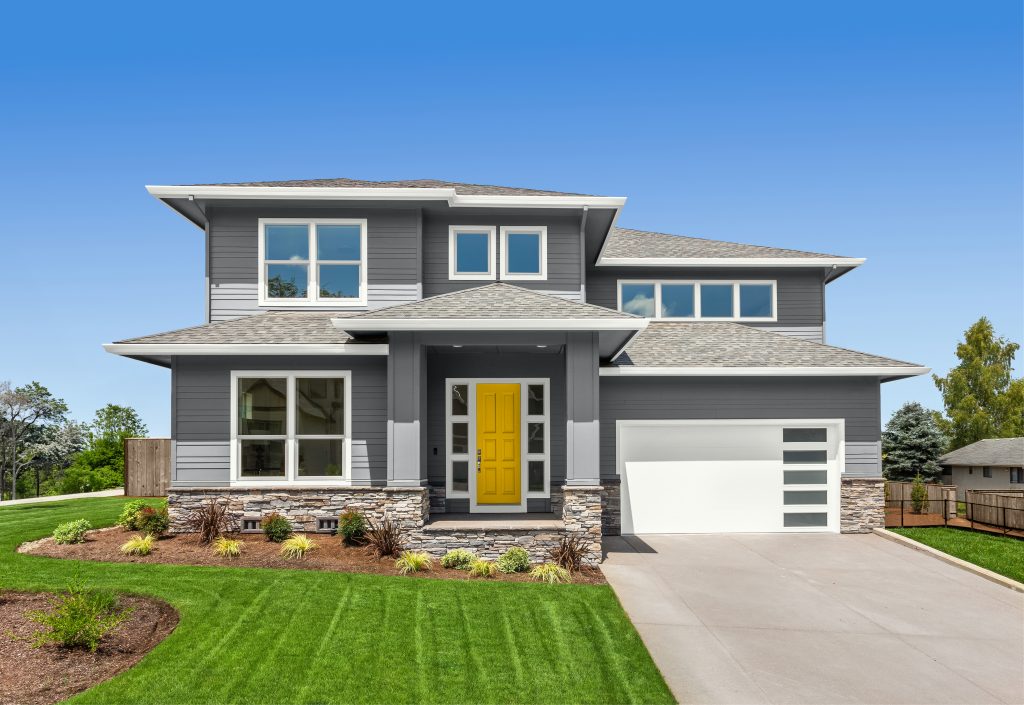 A new construction two-story gray house with a bright yellow front door, white trim, and a covered entry supported by square columns. The home features large windows, a stone-accent base, and an attached white garage under a clear blue sky. The wood fence has been finished with the 2026 BEHR wood stain color of the year, Taupe, in a semi-transparent finish. 