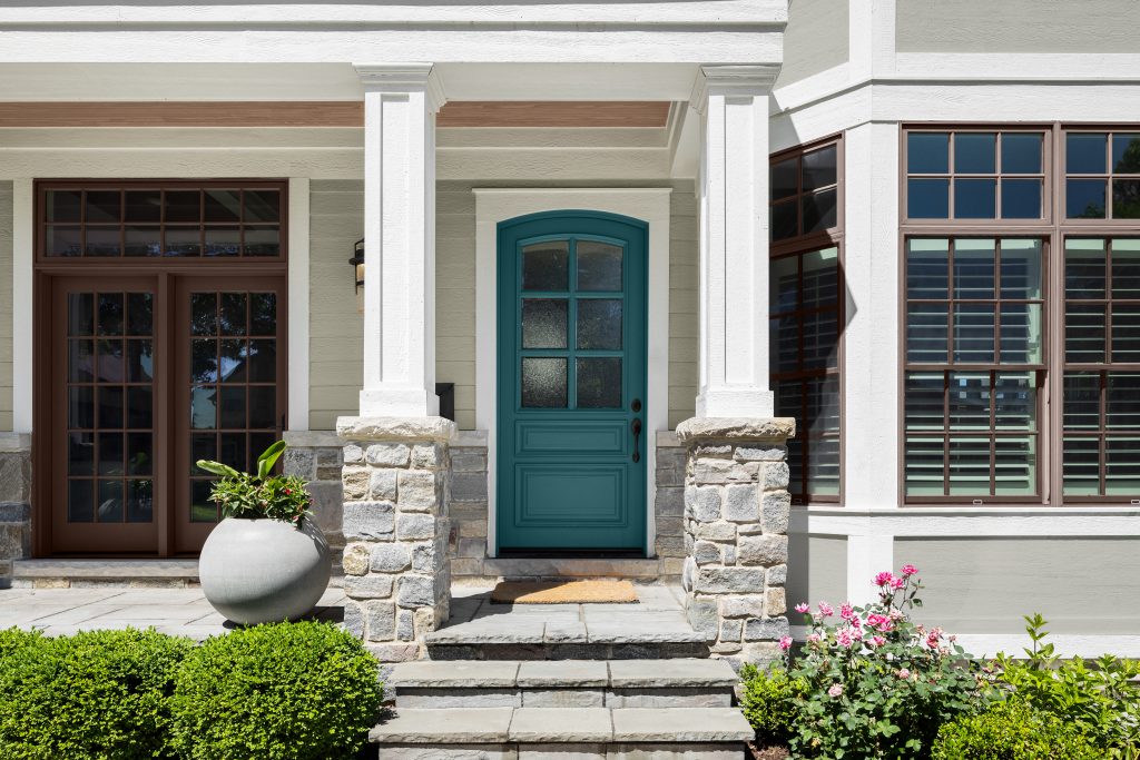 A front entry of a house with a deep sophisticated teal front door beneath a small, covered porch supported by white columns and stone bases. Stone steps lead up to the door, with a large red planter on the left and landscaped shrubs and pink flowers in the foreground. Large windows with dark trim frame the entrance.