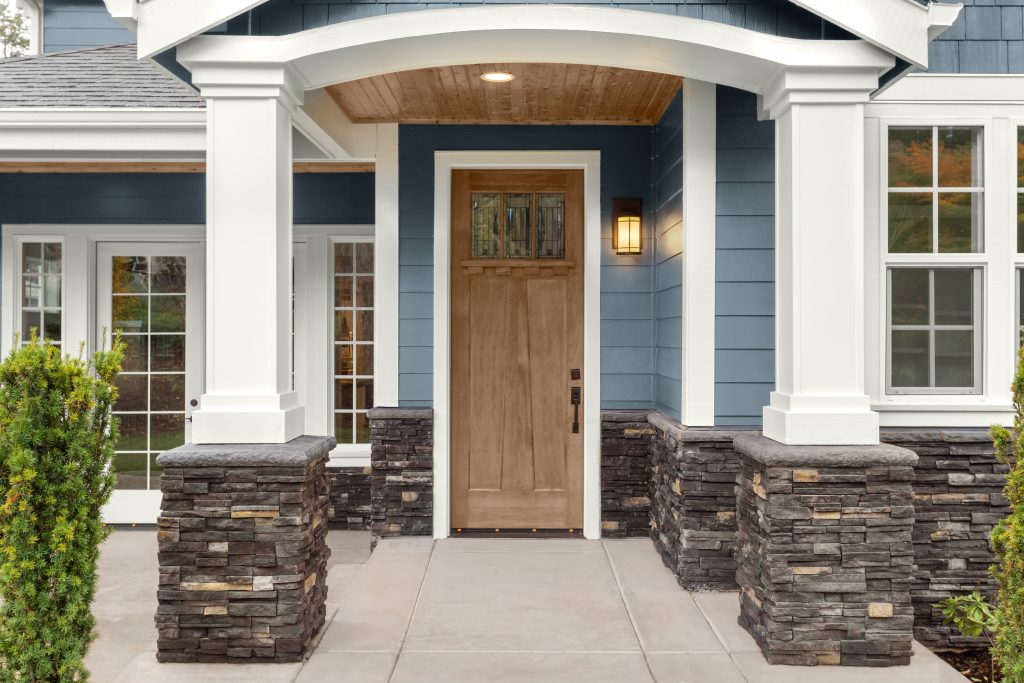 A front porch with a Taupe wood color entry door, blue siding, and stacked stone columns, featuring a wood-stained ceiling under the covered entry.