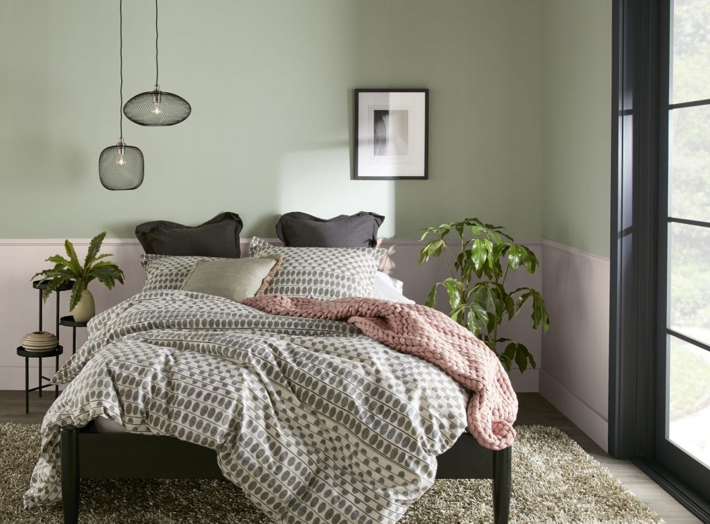 A serene bedroom with light pink and soft sage green walls and a dark bed frame dressed in patterned gray and white bedding. Layered pillows in neutral tones rest against the headboard, with a blush pink throw blanket draped across the bed. Two small pendant lights hang on one side, and a framed black-and-white artwork is centered above the bed.