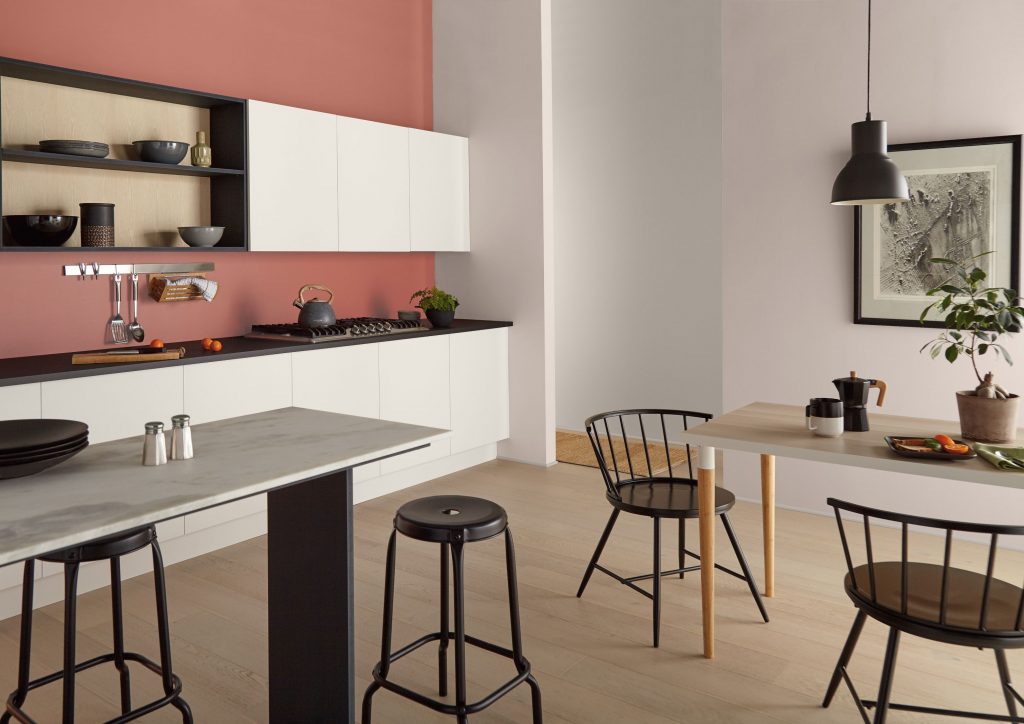 A modern kitchen and dining area with light pink walls and a terracotta accent wall above white upper cabinets and open wood shelving. A long gray countertop with black bar stools sits in the foreground, while a small light wood dining table with black wire chairs is placed nearby.