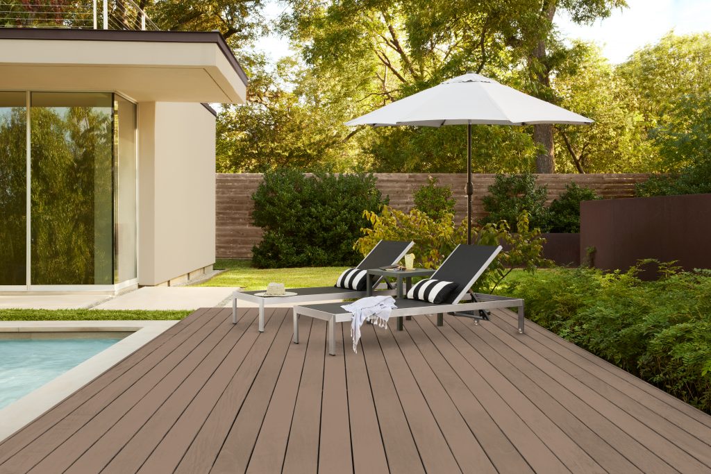 A modern backyard patio featuring a taupe-stained wood deck and matching taupe roof trim. The deck includes two lounge chairs, a white umbrella, and a small table beside a pool, all surrounded by lush greenery. The fence is stained in Baronial Brown, a previous Color of the Year.
