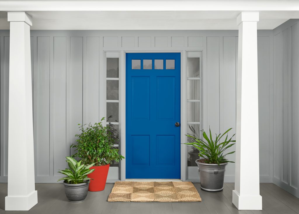 A front porch with light gray siding and white columns, featuring a bright blue front door centered between two narrow windows. Potted green plants sit on either side of the door, and a woven doormat rests on the gray floorboards.