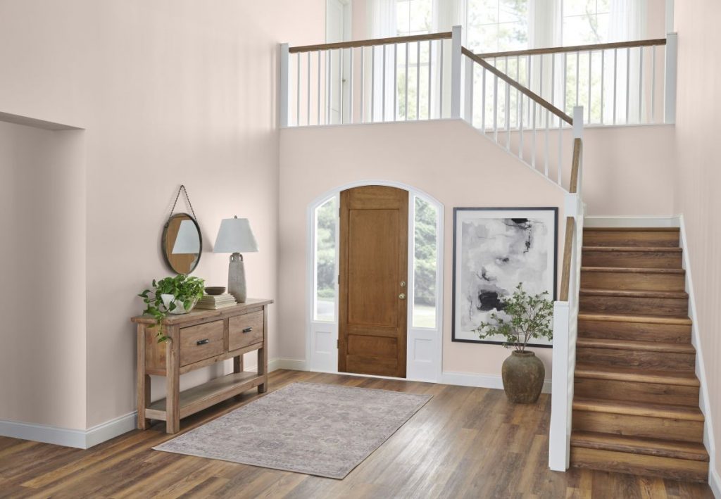 An airy high ceiling foyer with light pink walls, a wooden front door framed by arched windows, a staircase with walnut and white railing, a console table with lamp and plants, and a neutral area rug on wood floors.