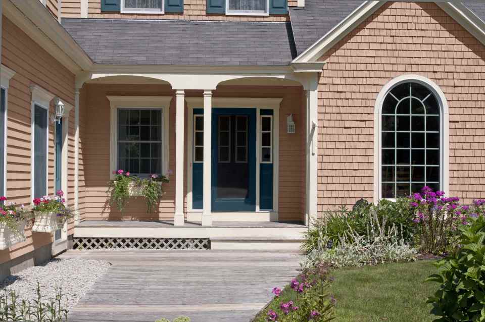A charming coastal-style home exterior with shingle siding painted Iced Copper, crisp Blank Canvas trim, and a bold Nocturne Blue front door, featuring a covered porch, white columns, and a landscaped walkway leading through a colorful garden.