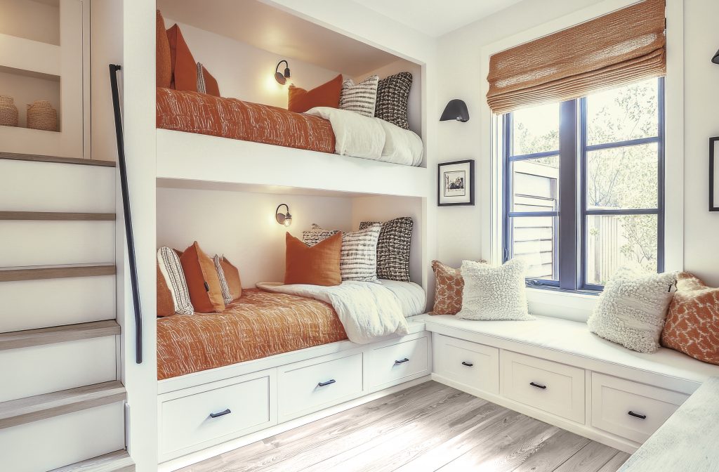 A bright, casual bedroom with built-in white bunk beds featuring orange and white bedding, including patterned pillows. A small ladder leads to the top bunk. A cozy window nook with cushioned seating and drawers underneath sits beside a large window with a woven shade. The room is accented with warm lighting, framed wall art, and an orange patterned rug on the floor.