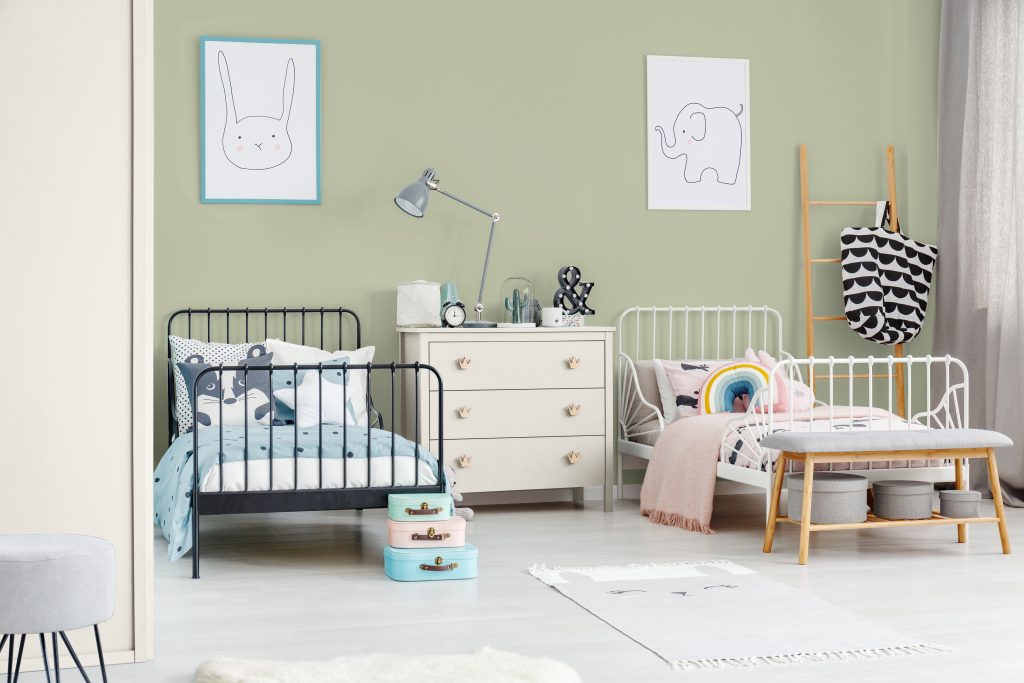 A shared children’s bedroom with a soft green accent wall, featuring two metal twin size beds, light wood furniture, blue and white and pink bedding, and playful animal artwork above the beds. 