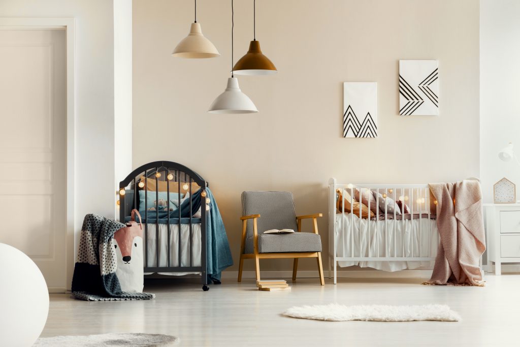 A stylish nursery with warm neutral walls, featuring two cribs with soft bedding, modern pendant lights in muted tones, cozy seating, minimal wall art, and layered textiles creating a calm, contemporary feel.