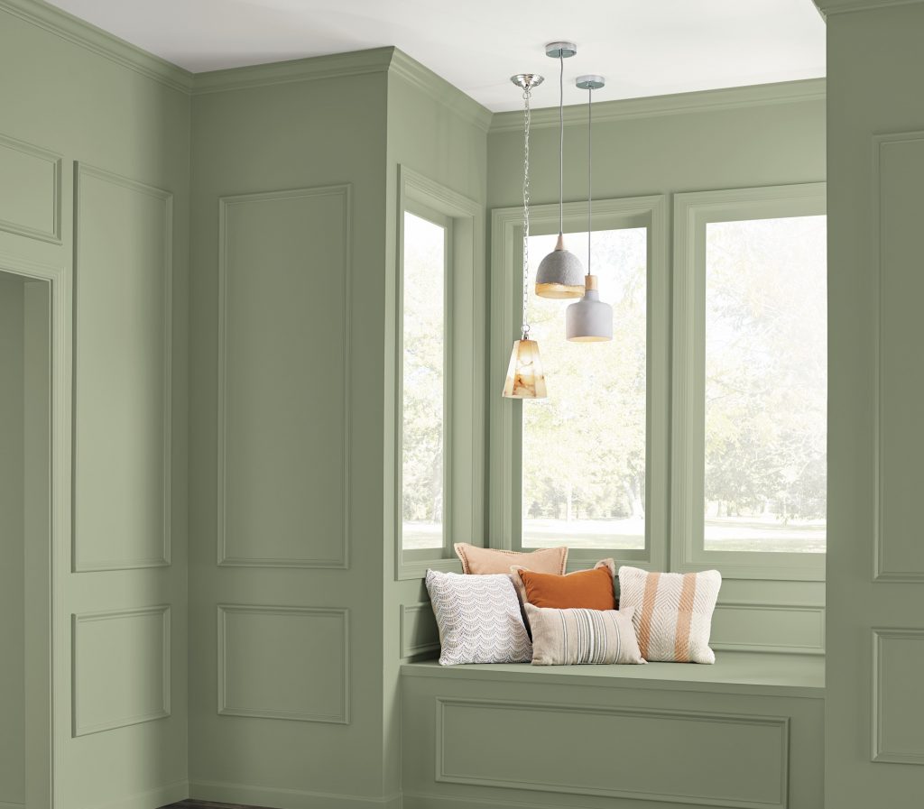 A cozy window nook with paneled walls painted in “Ecological,” featuring a built-in bench with neutral and warm-toned cushions under hanging pendant lights.