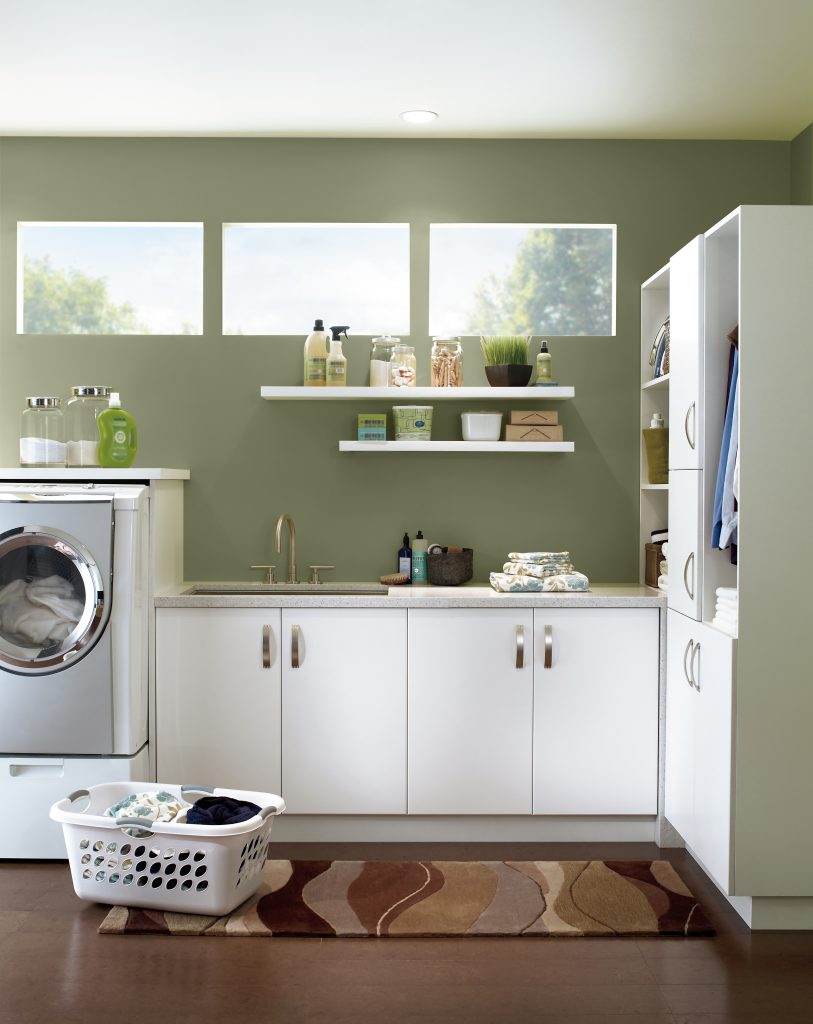 A green laundry room with walls painted in “Ecological,” featuring white cabinets, open shelving, a front-loading washer, and a basket of laundry for a clean, functional look.