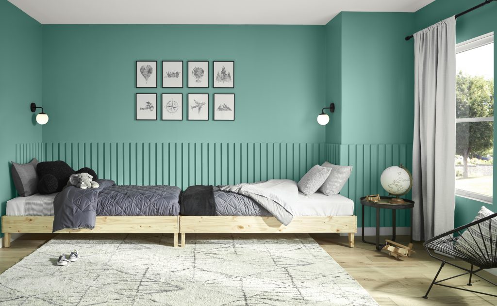 A modern kids’ bedroom with midtone green painted walls and paneled detail, featuring two low wooden twin beds with gray bedding, minimal black-and-white artwork above the beds, and natural light from a window creating a calm, cohesive space.
