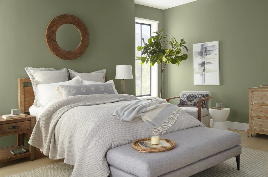 A relaxing green bedroom with walls painted in “Ecological,” featuring a light wood bed, layered neutral bedding, a cushioned bench, and soft natural accents.