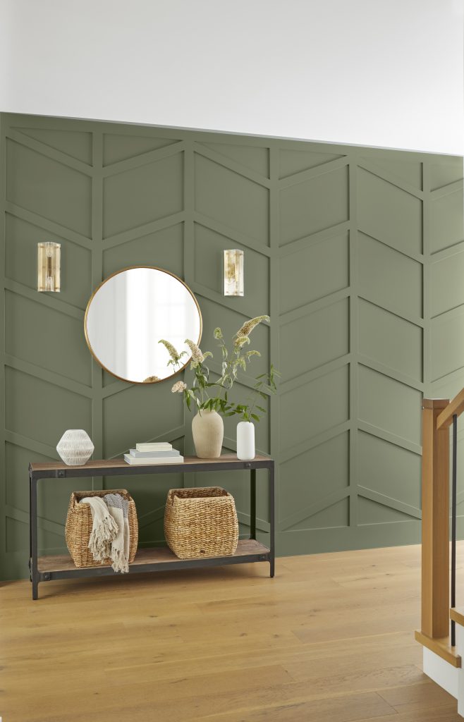 An entryway with geometric paneled walls painted in “Ecological,” featuring a round mirror, wall sconces, and a console table with woven baskets and decor.